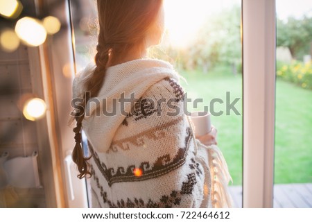Thoughtful young brunette woman with cup of coffee looking through the window, blurry garden outside