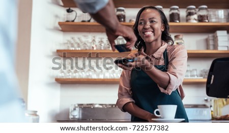 Smiling barista using nfs technology to help a customer pay for a purchase with their bank card in a cafe  Royalty-Free Stock Photo #722577382