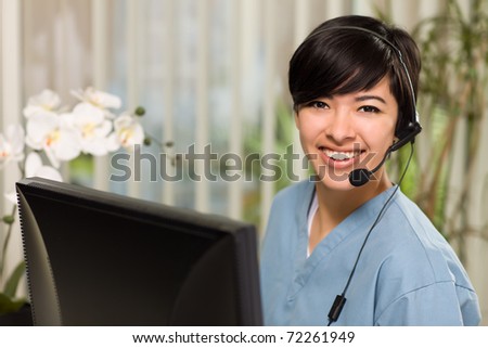 Smiling Attractive Multi-ethnic Young Woman Wearing Headset and Scrubs Near Her Computer Monitor. Royalty-Free Stock Photo #72261949