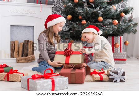Cute happy excited children, boy and girl in santa hats unwrap christmas present box in beautiful room. Sister and brother give Xmas gifts near decorated tree and fireplace. Family holiday