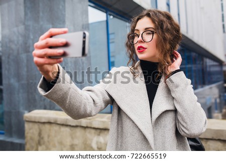young beautiful woman walking in the city street in grey coat, autumn fashion style, glasses, holding smartphone, taking selfie picture, smiling, happy
