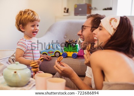 The young smiling parents sitting at the table in the room and eating bagels near little joyful son who looking at them