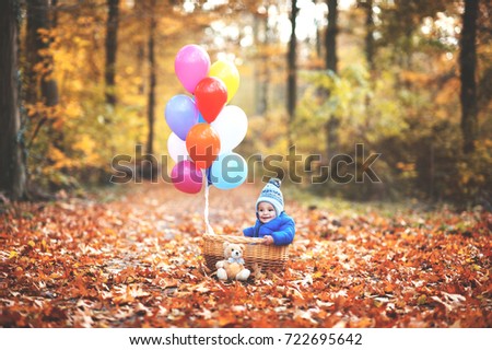 Little sweet boy is sitting in the basket with balloons which is going to fly into the air. He is in the forest which has autumn colors.
