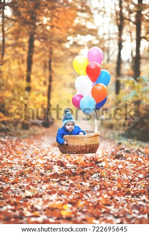 Little sweet boy is sitting in the basket with balloons which is flying into the air. He is in the forest which has autumn colors.