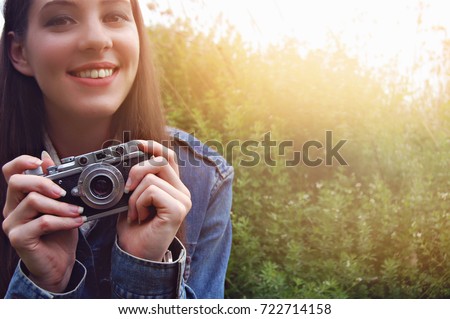 Young beautiful girl is smiling and looking at the viewer.  Pretty woman is holding retro old vintage camera in her hands. Female at nature green grass background in sunlight of  morning.