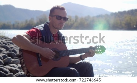 Man wearing sunglasses plays on a guitar sitting by mountain river on sunny day
