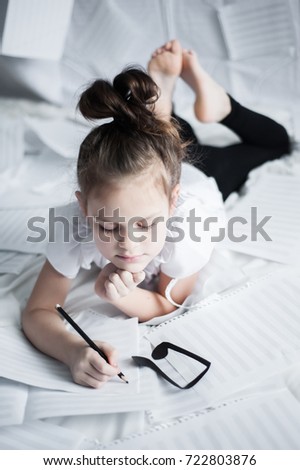 A little girl lies on her belly among clean sheets of paper to record musical notes next to a black note figure and holds a black pencil