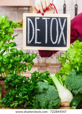 Young woman in kitchen having many green vegetables presenting board with detox sign.