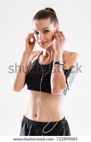 Portrait of a joyful fitness woman in sportswear listening to music with earphones while standing and looking at camera isolated over white background