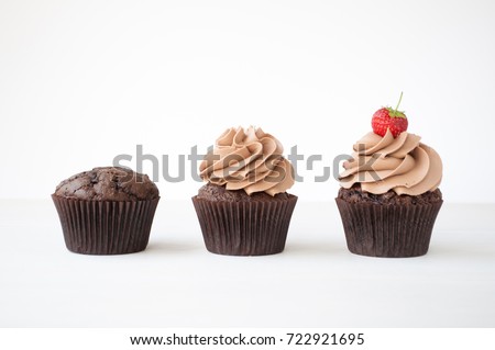 Cupcakes with whipped chocolate cream, decorated fresh strawberry on white wooden table. Picture for a menu or a confectionery catalog, showing different stages of creation.