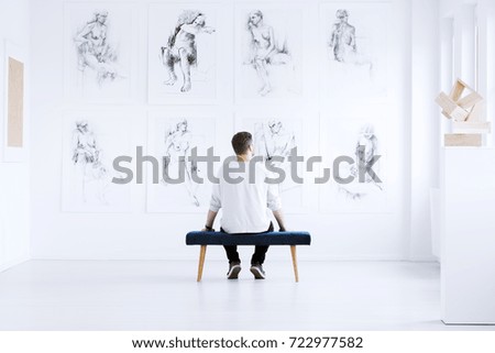 Man relaxing in art gallery while sitting on stool in front of white wall with drawings. Art gallery concept Royalty-Free Stock Photo #722977582