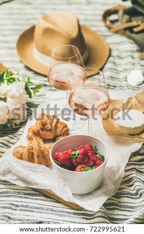 French style romantic summer picnic setting. Flat lay of glasses of rose wine with ice, strawberries in bowl, croissants, brie cheese, straw hat, peony flowers, square crop. Outdoor gathering concept