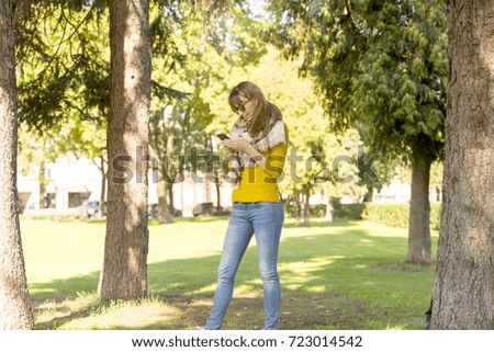 Cute woman is reading pleasant text message on mobile phone while taking a walk in the park in warm fall day