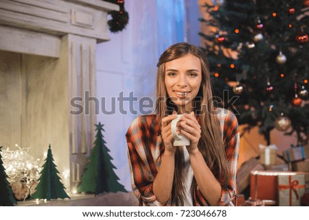 portrait of smiling woman with cup of hot drink in hands looking at camera in festive decorated room