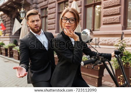 Displeased bearded business man sitting on modern motorbike outdoors with elegant woman which looking at mirror
