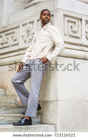 Portrait of Young Handsome African American Man in New York. Young black man wearing light color jacket, gray pants, black leather shoes, standing against vintage marble wall, looking around, relaxing