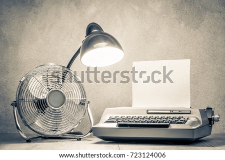 Retro typewriter with paper blank, old desk lamp and classic cooling fan on wooden table front concrete wall background. Vintage style sepia photo