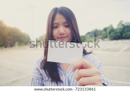 Image of a woman holding a blank piece of paper or card in white and she is smiling. Yellow flare. The background is a outdoor parking lot and concrete floor and tree.