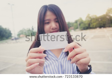 Image of a woman holding a blank piece of paper or card in white and she is smiling. Yellow flare. The background is a outdoor parking lot and concrete floor and tree.