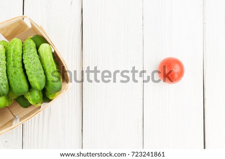 Lot of fresh green cucumbers in wicker basket and single red ripe tomato on old white wooden background