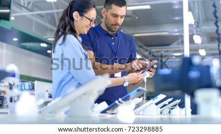 In the Electronics Store Consultant Gives Professional Advice to a Young Woman, She Considers Buying New Tablet Computer and Needs Expert Opinion. Store is Modern, Bright and Has all the New Devices. Royalty-Free Stock Photo #723298885