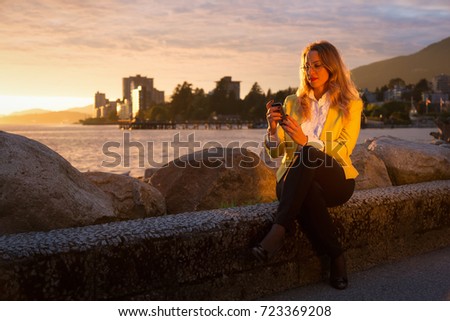 Well dressed professional and beautiful business woman is sitting and looking at her phone during a vibrant sunset. Picture taken in West Vancouver, British Columbia, Canada.
