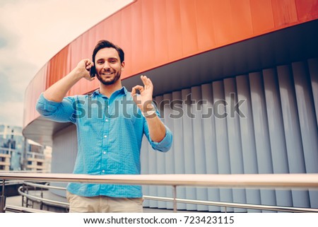 Half-length portrait of male tourist standing on urban setting making phone call in roaming enjoying international connection, happy hipster guy showing ok sign during mobile talk having cheap tariff