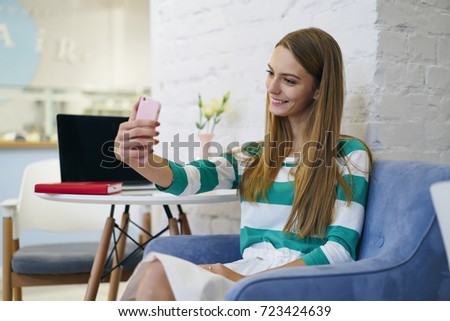 Charming young girl posing at camera while having coffee break in modern cafe interior, attractive female student making selfie for social networks updating profile photo using wifi connection