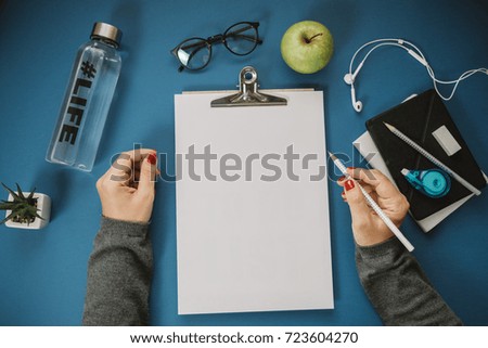 Woman hands with sheet of paper, pencils, water, apple, phone, glasses on a blue background. Top view, flat lay