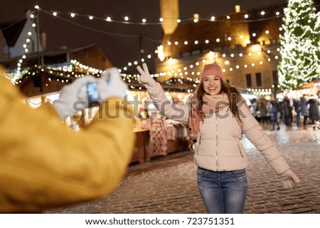 holidays, winter and people concept - happy woman posing for boyfriend with smartphone on square at christmas market