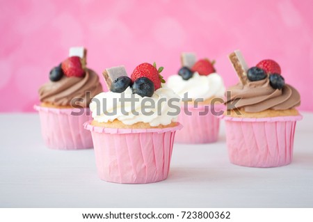 Pink cupcakes with white and chocolate whipped cream decorated with strawberry, blueberry, granola bar on white wooden table and pink background. Picture for a menu or a confectionery catalog.