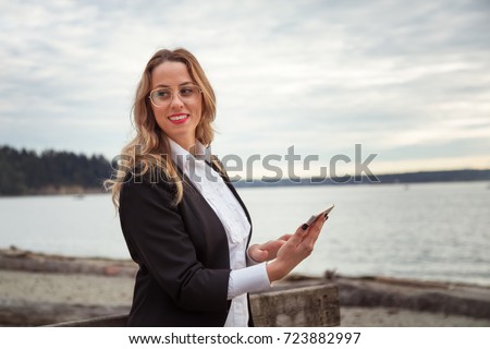 Young European Business Woman well dressed is holding her cell phone and smiling on a sandy beach. Picture taken West Vancouver, British Columbia, Canada. 