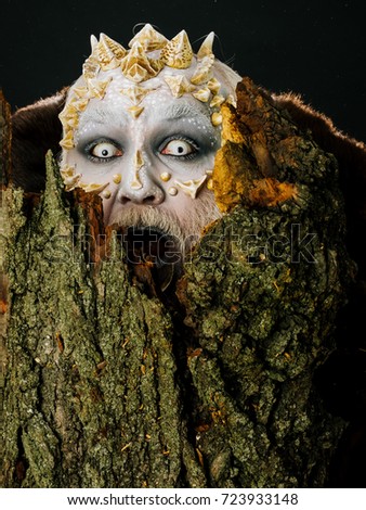 Goblin shouting with horns on head. Monster with sharp thorns and warts. Druid behind old bark on black background. Tree spirit and fantasy concept. Man with dragon skin and scary face.