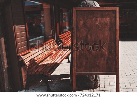 Mock-up of wooden black planking banner of chestnut colour staying on paving stone on summer sunny street with benches and glass-cases behind; empty wooden placeholder outdoors on bright day