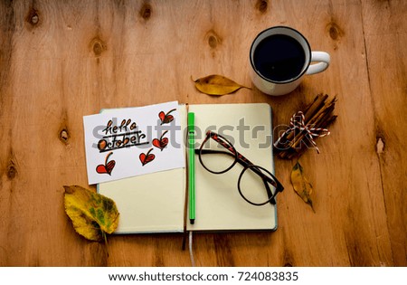 Coffee cup and an open notebook with hello October text and a pair of glasses and autumn leaves on rustic wooden table