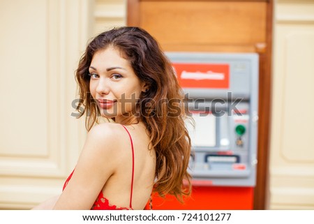 Young beautiful woman in a red dress stands against the background of an ATM in a shopping center