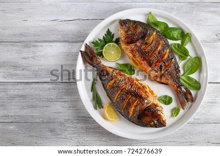 delicious grilled dorado or sea bream fish with lemon and orange slices, spices, fresh parsley and spinach on white platter on old wooden table, horizontal view from above Royalty-Free Stock Photo #724276639