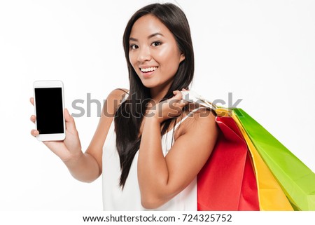 Portrait of a smiling young asian woman showing blank screen mobile phone while standing with colorful shopping bags isolated over white background