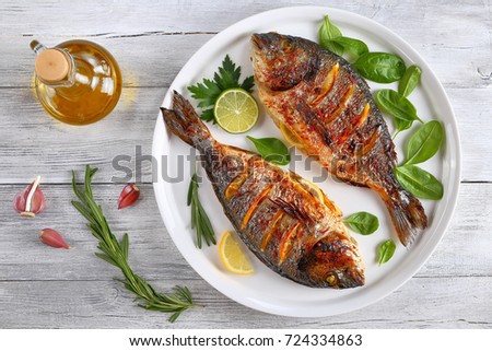roasted dorado or sea bream fish with lemon and orange slices, spices, fresh parsley and spinach on white platter on old wooden table with ingredients on background, horizontal view from above Royalty-Free Stock Photo #724334863