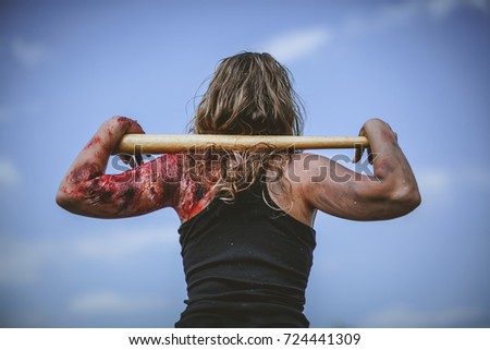 Blond girl in a black tank top and a burnt, bloody shoulder (practical special effect), looking up at the blue sky with a baseball bat in her hands