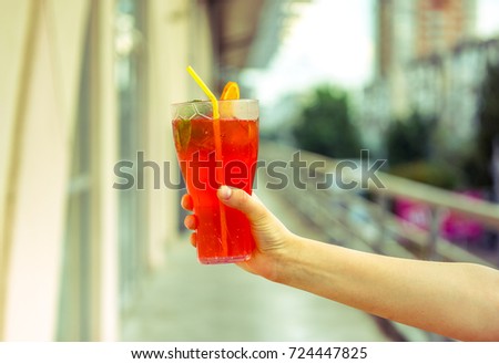 blurry effect on fresh cold red coral fruit watermelon strawberry mojito cocktail with mint, peace of ice, slice of orange and strong cocktail tube in strong man's hand against blurry city background.