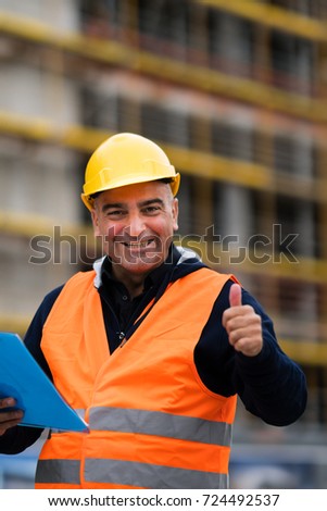 Cheerful and successful construction worker posing showing thumbs up gesture. Outdoors. Selective focus