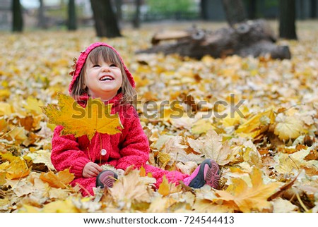 An autumn picture of a smiling little girl playing with yellow leaves in park. Baby wearing pink knitted overall
