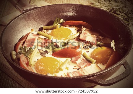 Bacon, eggs and tomatoes,  breakfast in a cast iron frying pan on a wooden board. Soft focus. Horizontal.