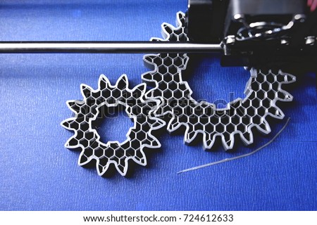 FDM 3D-printer manufacturing spur gears from silver-gray filament on blue print tape - top view on object, print bed and print head - matte look - foreground blanked out blurry