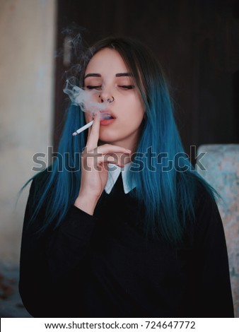 Emo girl smoking cigarette. Young student or pupil with blue colorful dyed hair, hat, piercing,lenses,ears tunnels and unusual hairstyle stands on black background.