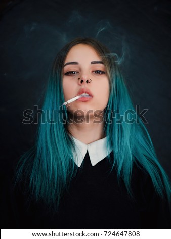 Emo girl smoking cigarette. Young student or pupil with blue colorful dyed hair, hat, piercing,lenses,ears tunnels and unusual hairstyle stands on black background.
