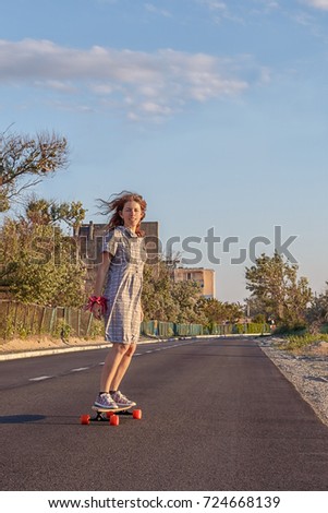 A girl in a dress with curly hair rides a long board on the asphalt.