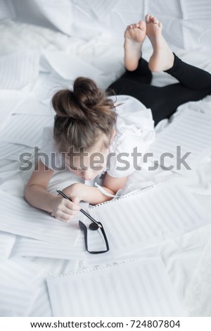 A little girl lies on her belly among blank sheets of paper to record musical notes and draws a black pencil a note model