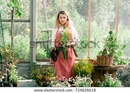 Young cute lady working at the greenhouse wearing pink linen apron, white gloves and leather belt with florist tools